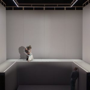 Eine große, helle Bühnenkonstruktion mit zwei Ebenen ist zu sehen. Oben links sitzt ein junger Mann, hinter ihm steht eine Frau; unten rechts steht ein Mann an der Wand. Die Bühne hat hohe graue Wände und leuchtende Linien an den Kanten.