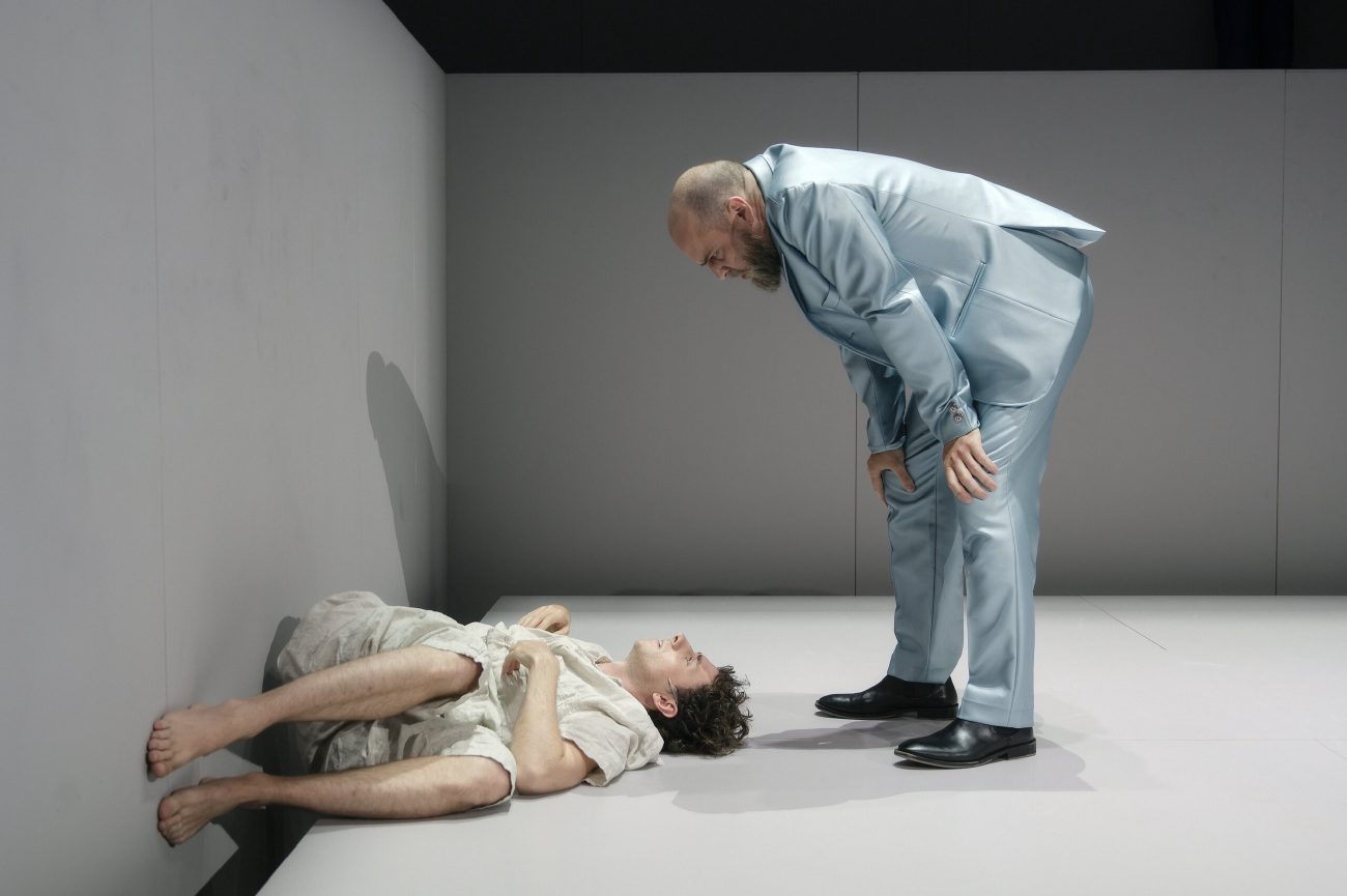 Ein junger Mann liegt auf einer hellen Bühnenkante und blickt zur Seite. Er trägt helle Kleidung und liegt seitlich mit angewinkelten Beinen. Auf der Wand und der Bühnenkante sind grünliche und violette Projektionen zu sehen.