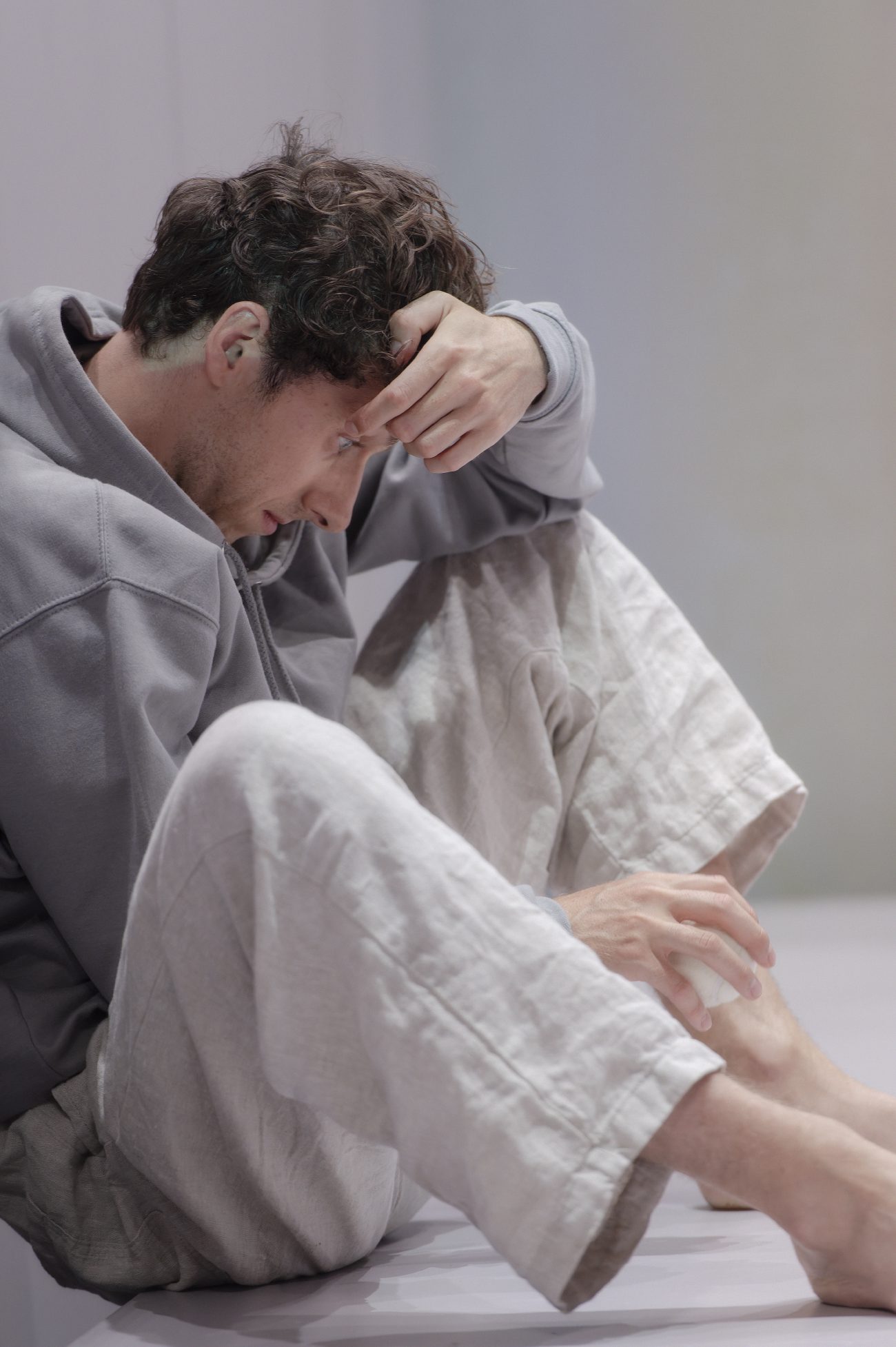 Eine rot beleuchtete Bühne zeigt oben eine Person in weißer Kleidung und unten einen jungen Mann, der mit dem Rücken zum Publikum sitzt. Die obere Person trägt eine Kappe und eine helle Arbeitskleidung und hält einen Gegenstand in der Hand. Der junge Mann sitzt auf dem Boden vor einer hohen Bühnenwand und trägt einen hellen Kapuzenpullover.