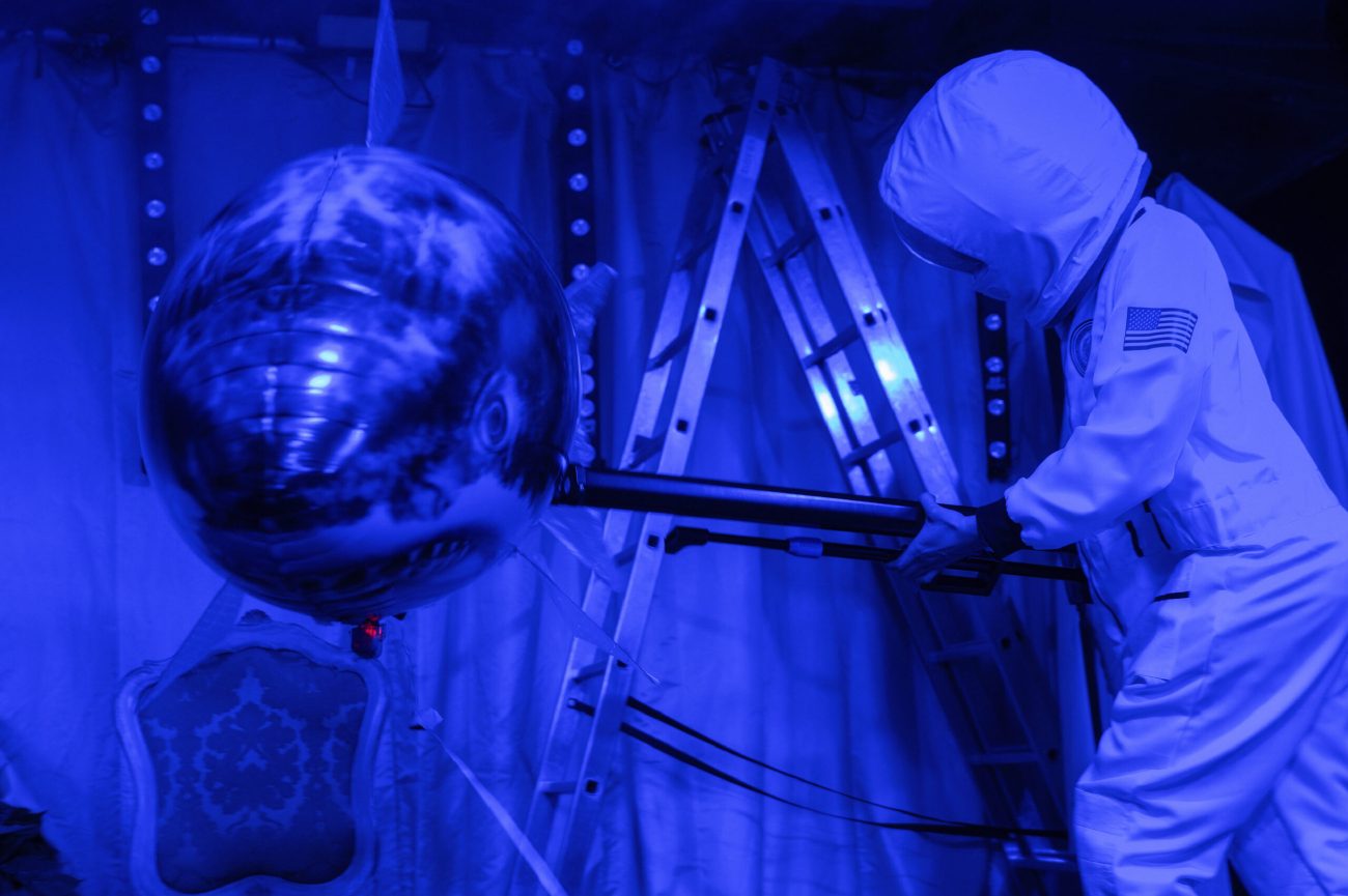 Eine Person in einem weißen Astronautenanzug mit Kapuze steht auf einer Bühne in blauem Licht. Sie hält eine Stange, an der ein großer Ballon in Form eines Haifischkopfs befestigt ist. Im Hintergrund sind Leitern, ein Vorhang und ein Stuhl zu sehen.