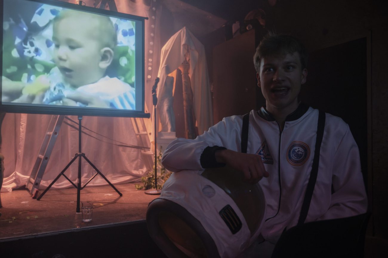 Ein junger Mann in einem weißen Astronautenanzug sitzt und hält einen Helm im Arm. Neben ihm steht eine Leinwand, auf der das Bild eines Babys mit einer gelben Spielfigur zu sehen ist. Im Hintergrund befinden sich eine Leiter, ein Mikrofonständer und abgedeckte Objekte.
