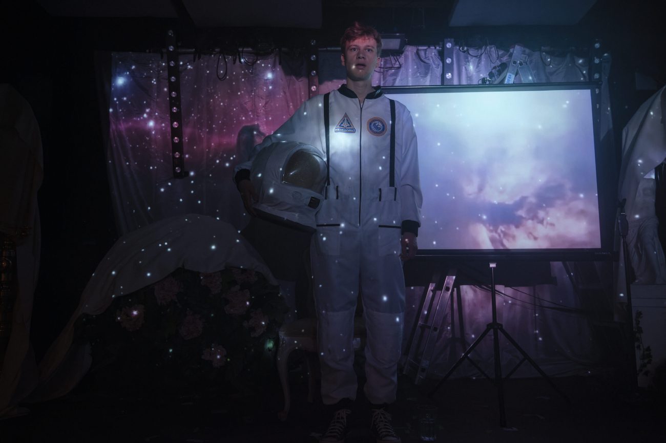 Ein junger Mann in einem weißen Astronautenanzug hält einen Helm unter dem Arm. Hinter ihm ist eine Leinwand mit einem Wolkenmotiv zu sehen, während im Raum Lichtpunkte wie Sterne projiziert werden. Links im Bild stehen Blumen und ein abgedeckter Gegenstand.