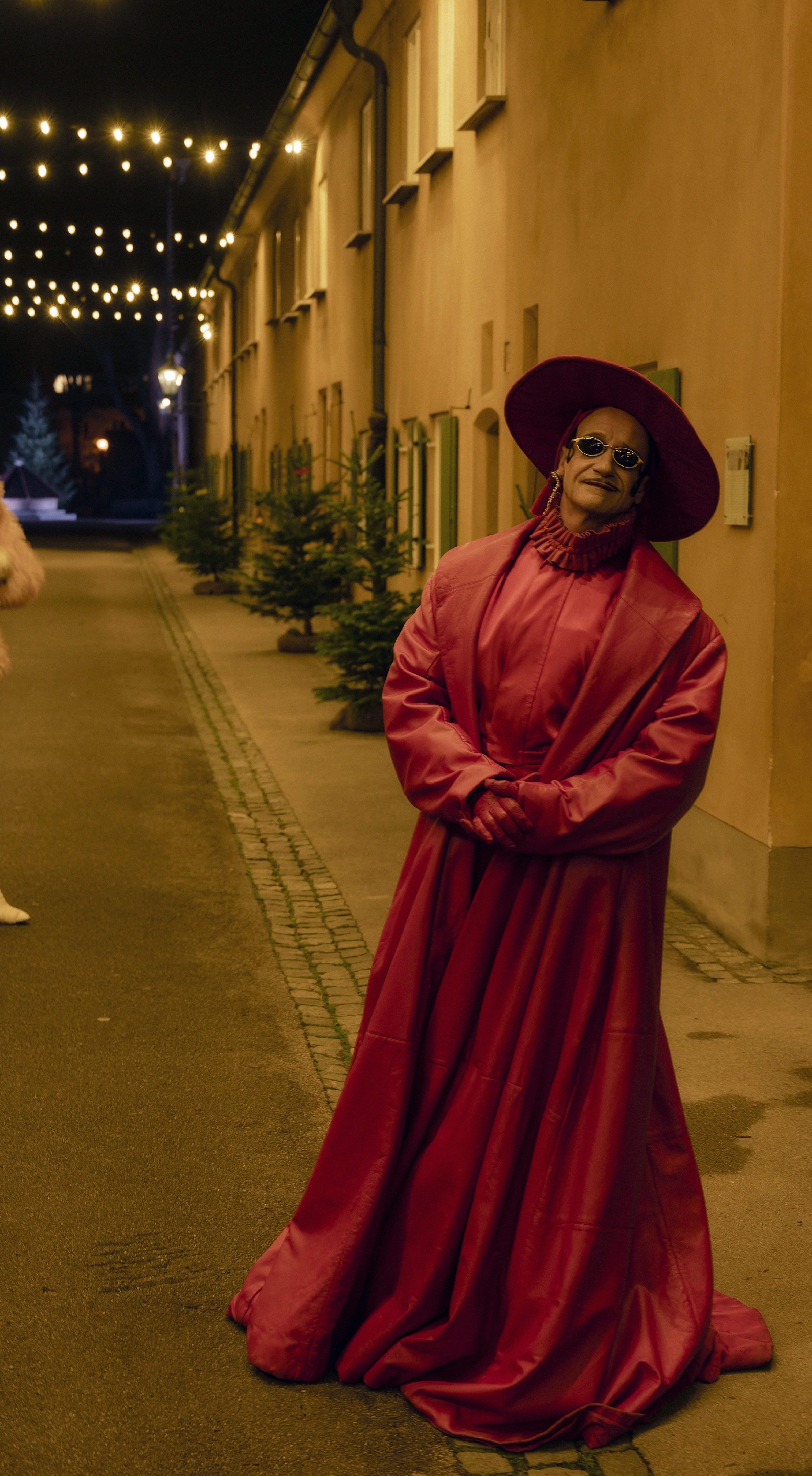 Zwei Personen stehen nachts in einer beleuchteten Straße zwischen Häusern. Vorne steht eine Person in einem langen roten Mantel mit Hut und Sonnenbrille, weiter hinten eine Person in rosa Kleidung mit Stiefeln. Über der Straße hängen Lichterketten, kleine Bäume stehen am Rand.