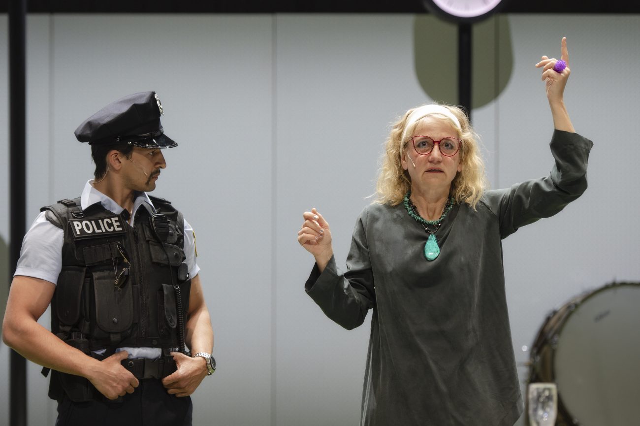 Zwei Frauen sitzen nebeneinander und schauen nach vorn. Die jüngere hat die Hände in den Schoß gelegt und trägt ein schwarzweiß-kariertes Kleid. Die ältere trägt eine rote Brille, lächelt und hat einen Arm mit geöffneter Hand über den Kopf gehoben.