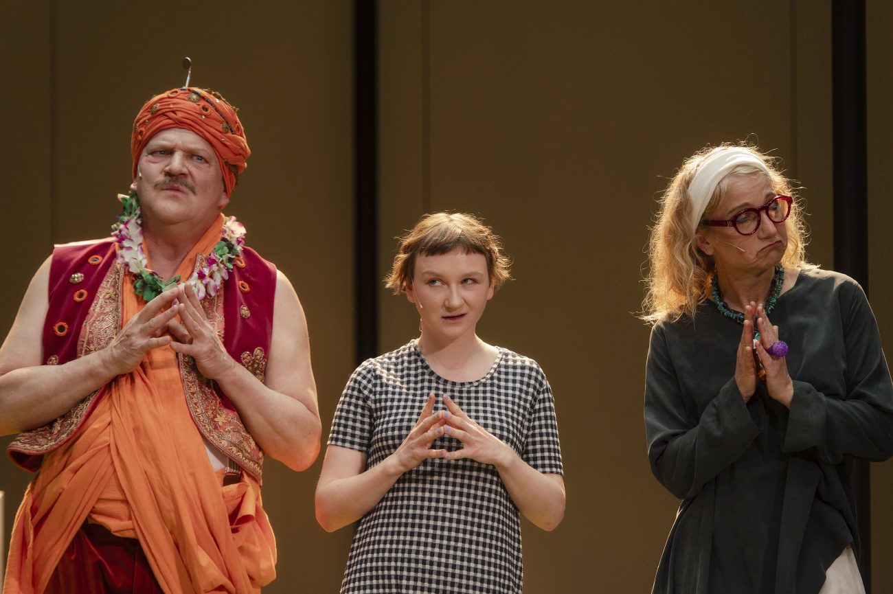 Ein Raum mit Bahnhofsuhren, Wänden und Rollrasen. Vorne rechts ein Mann in einem orangefarbenen indischen Kostüm, mit Schnurrbart und Turban, der beide Arme weit nach oben streckt, um ihn herum andere Personen, die ihn betrachten.